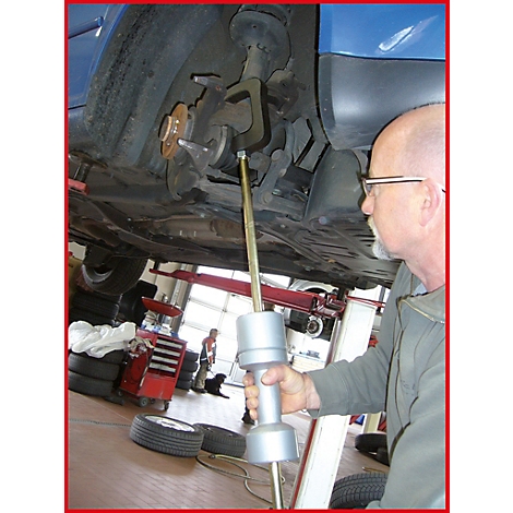 Ein Mechaniker inspiziert das Fahrwerk eines Autos. Er hält ein Werkzeug und steht unter dem Fahrzeug.